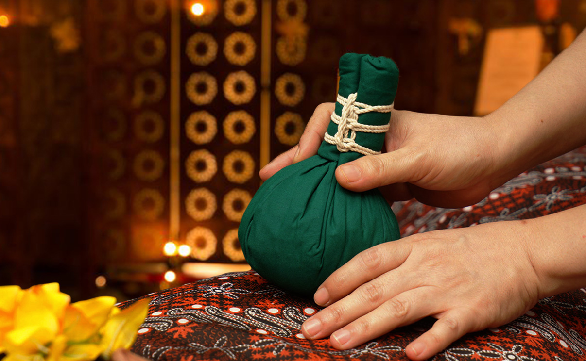 Close-up of hands holding a green herbal compress ball on patterned fabric, warm ambient lighting with candles adding a calm mood