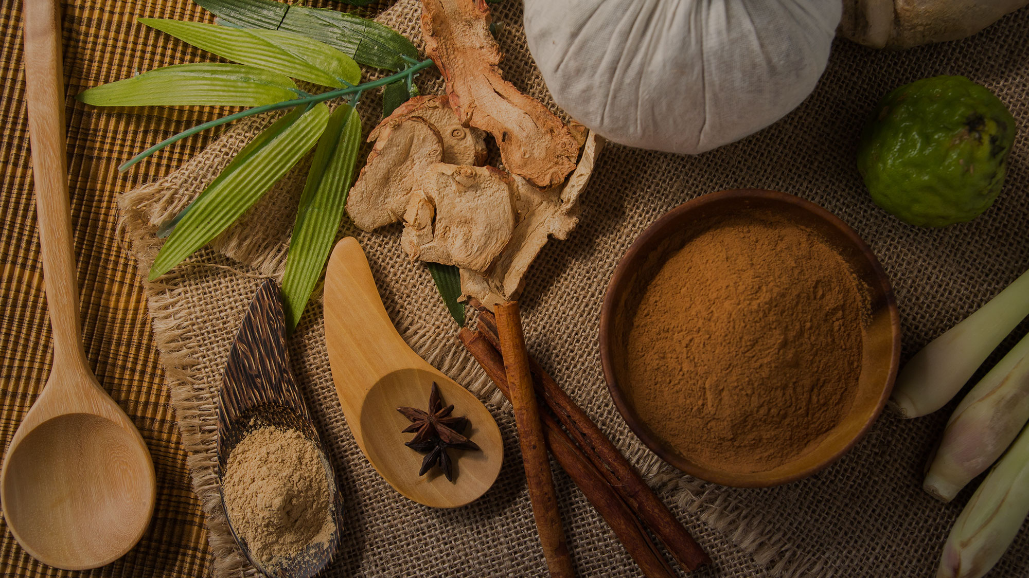 Overhead flat lay of spa ingredients including dried herbs, cinnamon sticks, cinnamon powder in a bowl, star anise, and wooden elements